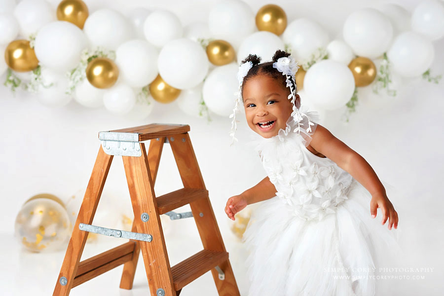 baby photographer near Carrollton, GA; girl laughing with ladder during white and gold studio milestone session